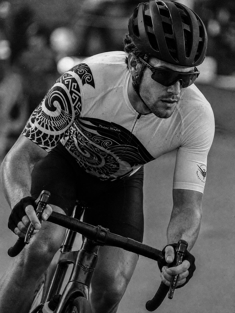 A black and white close-up of a cyclist in a white jersey with a large black Maori-style tribal graphic on the sleeve and chest, wearing a dark helmet and sunglasses while riding a road bike.